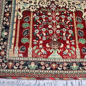 May include: A red prayer rug with a floral design and a central tree motif. The rug has a black and white border with a fringe.