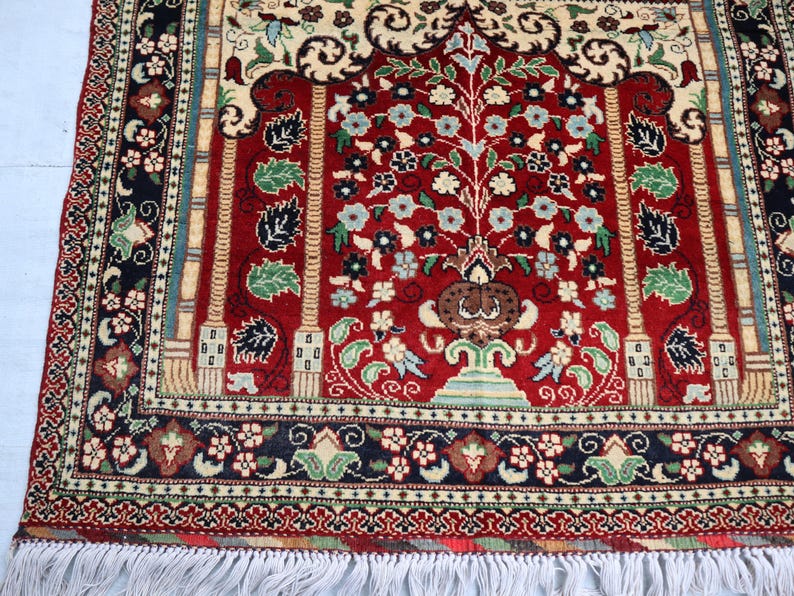 May include: A red prayer rug with a floral design and a central tree motif. The rug has a black and white border with a fringe.