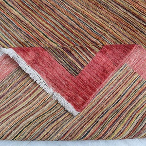 May include: Close-up of a multi-colored striped rug with a red border and white fringe. The rug is folded in half, showing the underside.