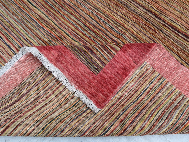 May include: Close-up of a multi-colored striped rug with a red border and white fringe. The rug is folded in half, showing the underside.