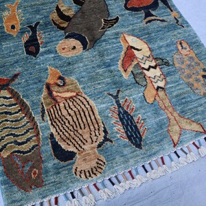 May include: A blue rug with a fringe border features a repeating pattern of stylized fish in various colors and designs. The rug has a vintage, hand-woven aesthetic.