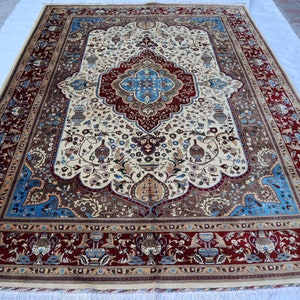 Puede incluir: Una alfombra persa tradicional con fondo crema y intrincados patrones florales en tonos rojos, azules y marrones. La alfombra tiene un borde con un diseño repetitivo de flores y hojas estilizadas.