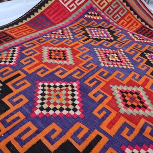 May include: A colourful, hand-woven rug with a geometric design. The rug features a rich palette of purple, orange, red, black, and white. The intricate patterns include squares and repeating geometric shapes, creating a visually striking textile.