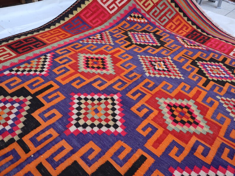 May include: A colourful, hand-woven rug with a geometric design. The rug features a rich palette of purple, orange, red, black, and white. The intricate patterns include squares and repeating geometric shapes, creating a visually striking textile.