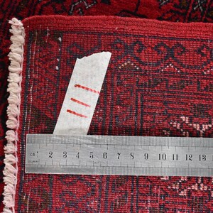 May include: Close-up of a red oriental rug with a detailed pattern. A metal ruler, marked in centimeters, is placed across the rug. The rug's edge features a fringe. The rug's pattern is dark red and black.