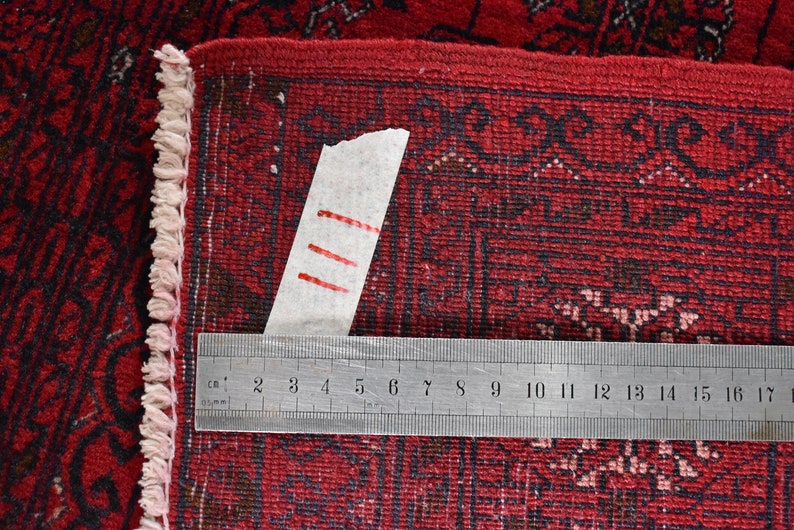 May include: Close-up of a red oriental rug with a detailed pattern. A metal ruler, marked in centimeters, is placed across the rug. The rug's edge features a fringe. The rug's pattern is dark red and black.