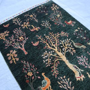 May include: A dark green rug with a floral pattern featuring trees, birds, and other designs. The rug has a fringe border.