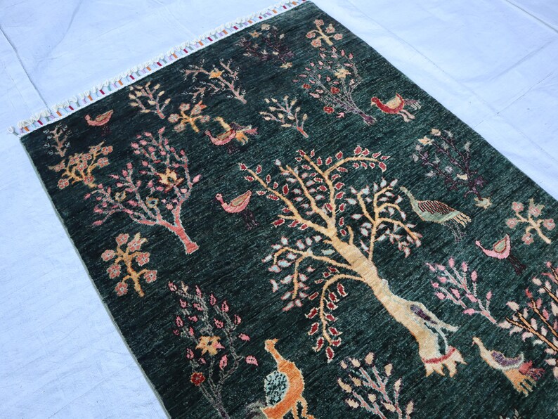 May include: A dark green rug with a floral pattern featuring trees, birds, and other designs. The rug has a fringe border.