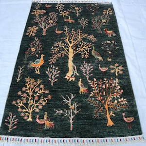 May include: A green rug with a floral and bird design. The rug features a central tree with branches and leaves, surrounded by smaller trees and birds. The rug has a fringe border.