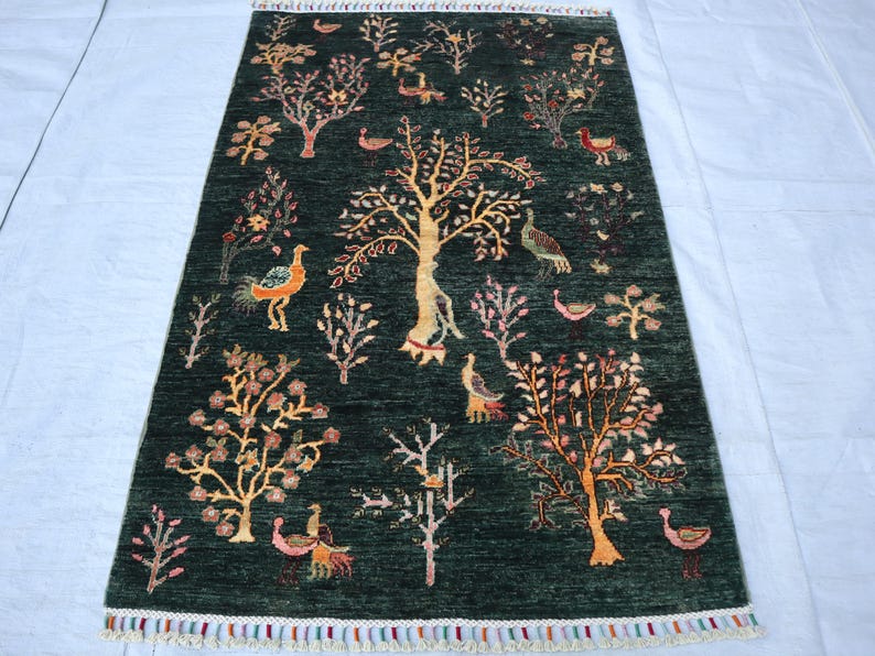 May include: A green rug with a floral and bird design. The rug features a central tree with branches and leaves, surrounded by smaller trees and birds. The rug has a fringe border.