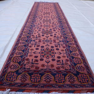 May include: A long rectangular runner rug featuring a dusty pink central field with repeating geometric floral motifs in dark blue, red, and yellow. The rug is framed by a wide dark blue border with stylized botanical patterns and white fringe.
