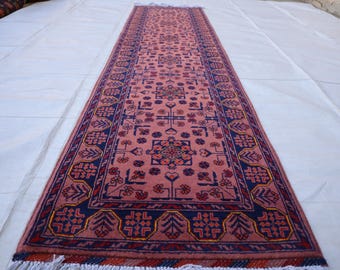 May include: A long rectangular runner rug featuring a dusty pink central field with repeating geometric floral motifs in dark blue, red, and yellow. The rug is framed by a wide dark blue border with stylized botanical patterns and white fringe.
