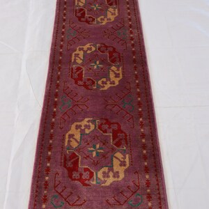 May include: A long, narrow rug with a pink background and intricate geometric patterns in shades of red, yellow, and green. The rug has a fringe border.