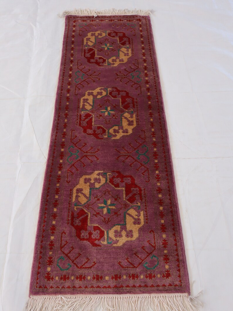 May include: A long, narrow rug with a pink background and intricate geometric patterns in shades of red, yellow, and green. The rug has a fringe border.