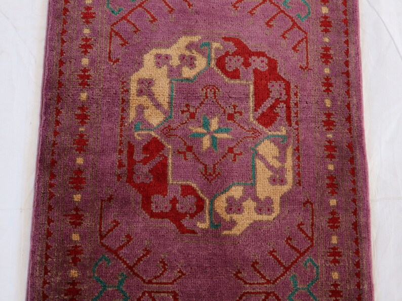 May include: A vintage rug with a purple background and a central design featuring red, cream, and teal accents. The rug has a geometric pattern with intricate details.