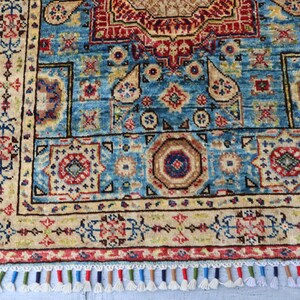May include: A detailed, rectangular rug with a central blue field, adorned with intricate geometric patterns in red, beige, and brown. The rug features a border with floral motifs and a fringe of colorful tassels. The rug's dimensions are not visible.