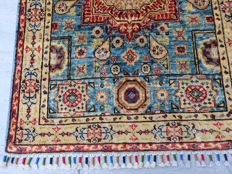 May include: A detailed, rectangular rug with a central blue field, adorned with intricate geometric patterns in red, beige, and brown. The rug features a border with floral motifs and a fringe of colorful tassels. The rug's dimensions are not visible.