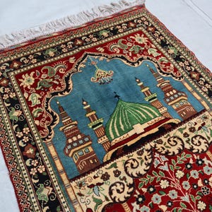 May include: A traditional prayer rug with a blue background and a central design of a mosque with a green dome. The rug has a red border with intricate floral patterns.