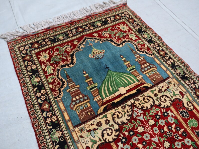 May include: A traditional prayer rug with a blue background and a central design of a mosque with a green dome. The rug has a red border with intricate floral patterns.