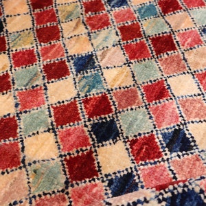May include: A close-up of a colorful checkered rug with red, pink, blue, and cream squares. The rug has a textured, woven look.