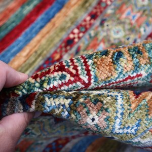 May include: Close-up of a colorful rug with intricate patterns in shades of red, blue, green, and beige. The rug is woven with a high-density pile, creating a luxurious and textured surface.
