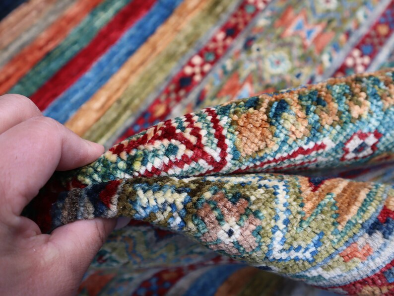 May include: Close-up of a colorful rug with intricate patterns in shades of red, blue, green, and beige. The rug is woven with a high-density pile, creating a luxurious and textured surface.