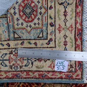 May include: Close-up of a section of a patterned rug with a ruler laid across it. The rug features a beige background with intricate designs in red, blue, and green. The edge has a fringe with colorful tassels. The tag reads "345/5.5".