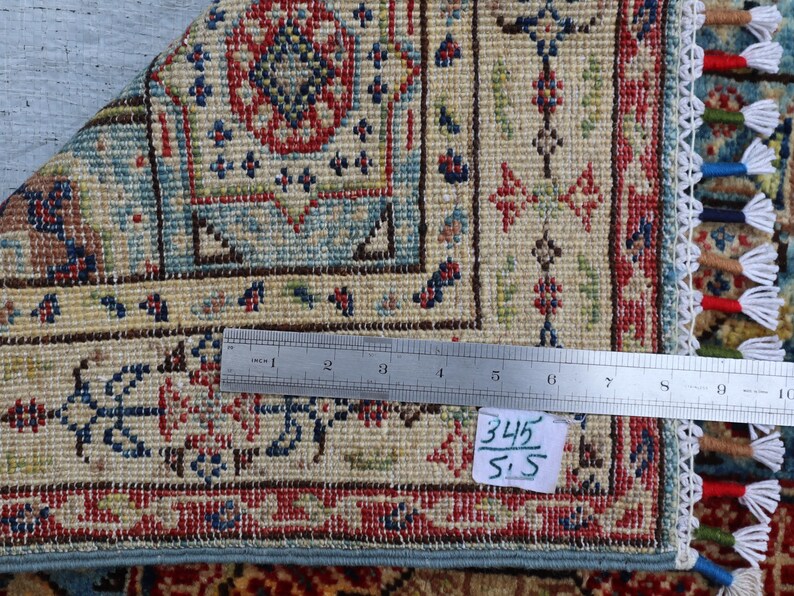 May include: Close-up of a section of a patterned rug with a ruler laid across it. The rug features a beige background with intricate designs in red, blue, and green. The edge has a fringe with colorful tassels. The tag reads "345/5.5".