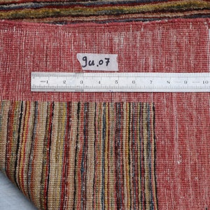May include: Close-up of the back of a red and multi-colored striped rug with a white fringe. A ruler is placed on the rug with the text "Ju.07" written on a piece of paper.