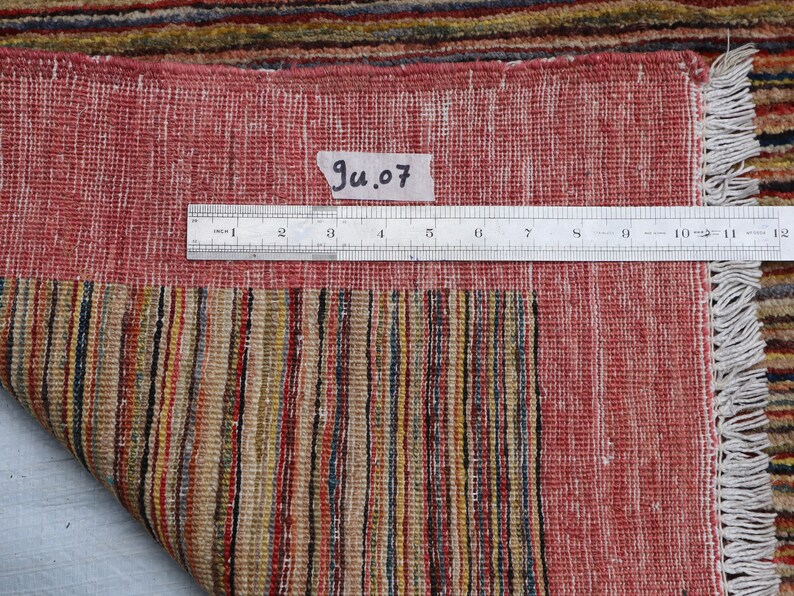 May include: Close-up of the back of a red and multi-colored striped rug with a white fringe. A ruler is placed on the rug with the text "Ju.07" written on a piece of paper.