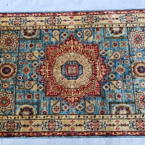 May include: Rectangular area rug with a central medallion design in shades of red, beige, and blue. The rug features an intricate pattern with floral and geometric motifs, and a fringed edge. The rug's border is a contrasting beige color.