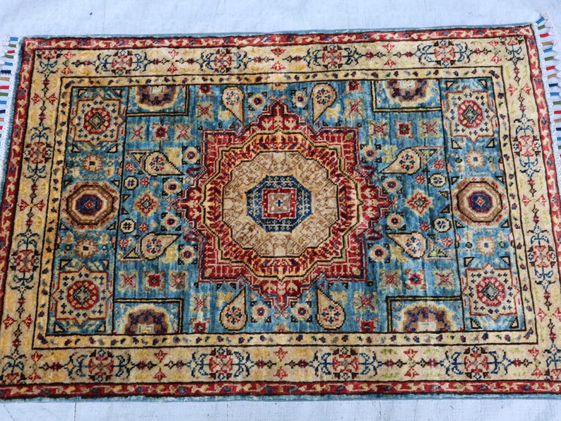May include: Rectangular area rug with a central medallion design in shades of red, beige, and blue. The rug features an intricate pattern with floral and geometric motifs, and a fringed edge. The rug's border is a contrasting beige color.