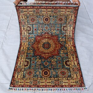 May include: A rectangular rug with a central medallion design in shades of red, gold, and blue. The rug features intricate floral and geometric patterns, with a cream-colored border and fringed edges. The rug is held up by unseen hands.