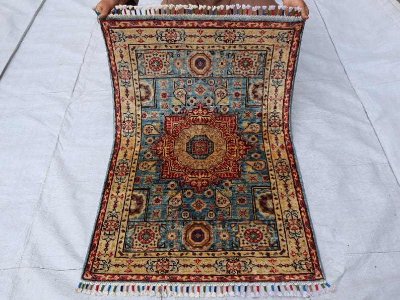 May include: A rectangular rug with a central medallion design in shades of red, gold, and blue. The rug features intricate floral and geometric patterns, with a cream-colored border and fringed edges. The rug is held up by unseen hands.