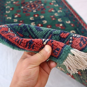 Pu&ograve; includere: Un tappeto verde, rosso e blu con frange. Il tappeto &egrave; piegato a met&agrave; ed &egrave; tenuto da una mano.