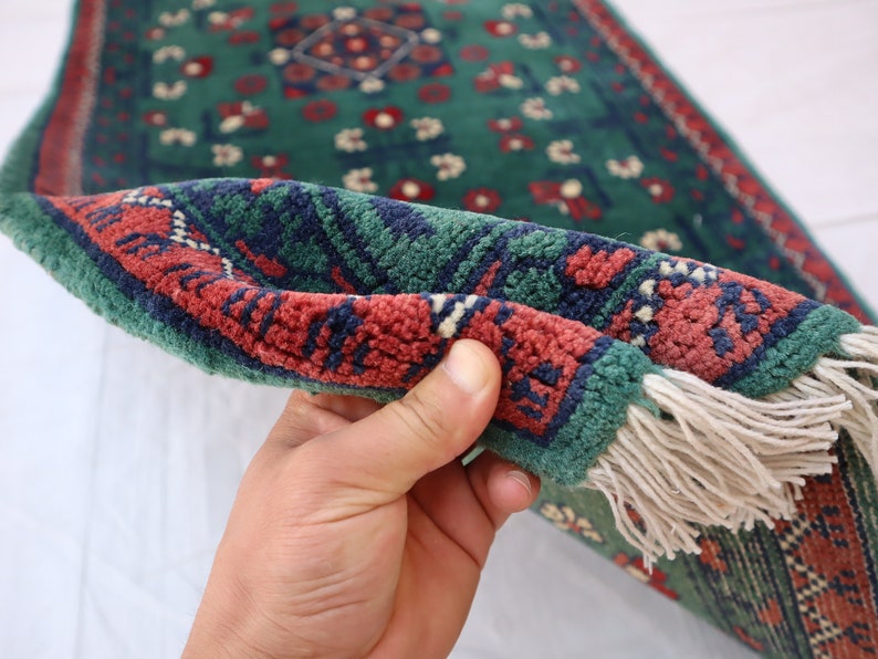 Pu&ograve; includere: Un tappeto verde, rosso e blu con frange. Il tappeto &egrave; piegato a met&agrave; ed &egrave; tenuto da una mano.
