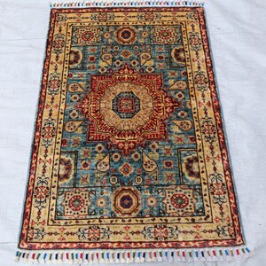 May include: Rectangular rug with an intricate, symmetrical design. The rug features a central medallion in shades of red and tan, surrounded by a teal background and a border of gold and red. The rug has a fringe of colorful tassels.