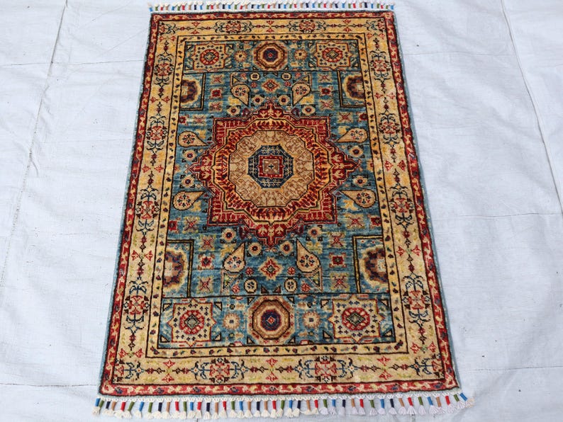 May include: Rectangular rug with an intricate, symmetrical design. The rug features a central medallion in shades of red and tan, surrounded by a teal background and a border of gold and red. The rug has a fringe of colorful tassels.