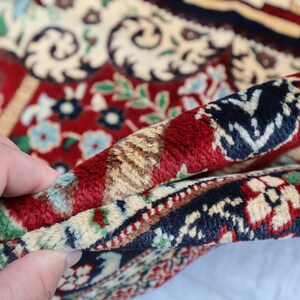 May include: Close-up of a red, cream and blue patterned rug with a hand holding the edge of the rug.