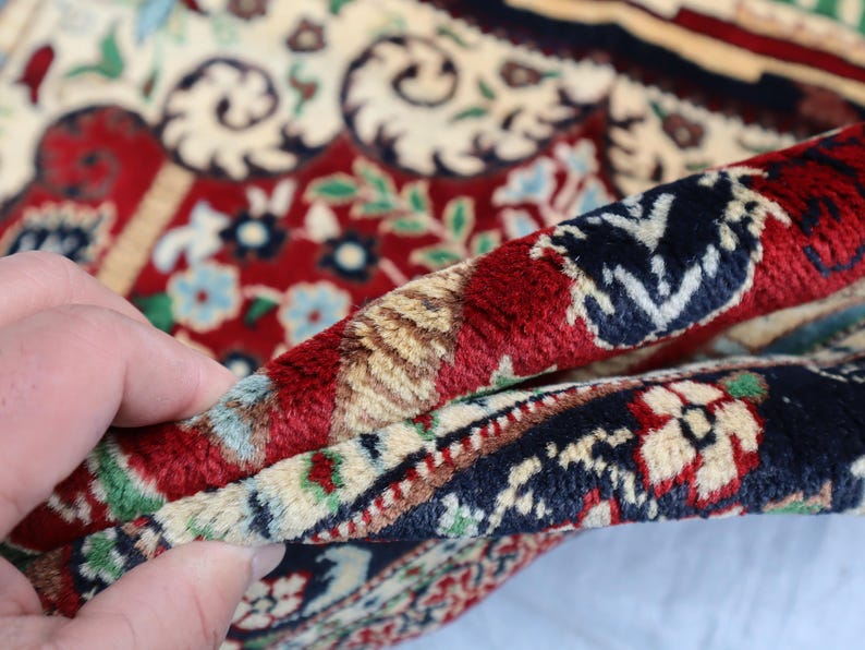 May include: Close-up of a red, cream and blue patterned rug with a hand holding the edge of the rug.