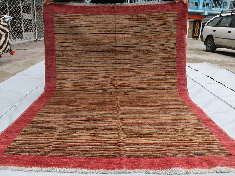 May include: A red and brown striped rug with a red border and fringe. The rug is folded in half and is being held by a person's hands.