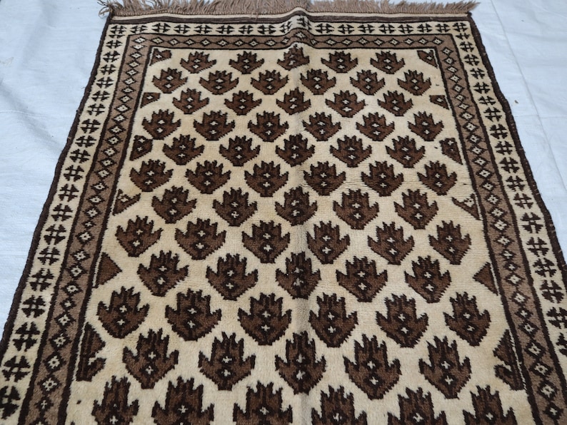 May include: A cream-colored rug with a brown geometric pattern. The pattern features repeating stylized flower shapes.