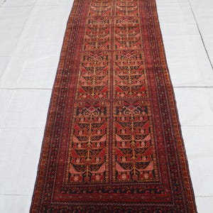 Könnte beinhalten: Ein langer, rechteckiger Teppich mit einem detaillierten Muster in Rot-, Braun- und Goldtönen. Das zentrale Design zeigt sich wiederholende florale oder baumartige Motive. Der Teppich hat einen dunklen Rand und gefranste Kanten und zeigt einen traditionellen Stil.