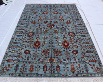 May include: A rectangular, light blue rug with an intricate floral pattern in shades of red, brown, and blue. The rug features a detailed border and a fringe along the edges. The design includes stylized flowers and botanical elements.