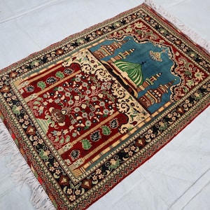 May include: A red, blue and green prayer rug with intricate floral and geometric patterns. The rug features a mosque design in the centre.