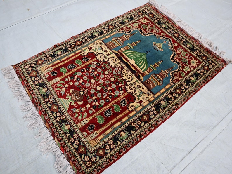 May include: A red, blue and green prayer rug with intricate floral and geometric patterns. The rug features a mosque design in the centre.