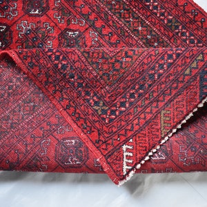 May include: A red oriental rug with a geometric pattern is partially rolled up, revealing the underside. The rug features a repeating design in black, brown, and white, with a fringed edge. The rug is on a white surface.