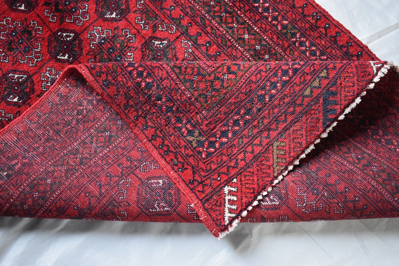 May include: A red oriental rug with a geometric pattern is partially rolled up, revealing the underside. The rug features a repeating design in black, brown, and white, with a fringed edge. The rug is on a white surface.
