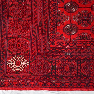 May include: Close-up of a vibrant red rug with intricate geometric patterns. The rug features a detailed border with repeating floral and geometric motifs in dark blue and cream. The central field showcases a symmetrical design with medallions and stylized shapes.