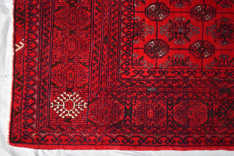 May include: Close-up of a vibrant red rug with intricate geometric patterns. The rug features a detailed border with repeating floral and geometric motifs in dark blue and cream. The central field showcases a symmetrical design with medallions and stylized shapes.